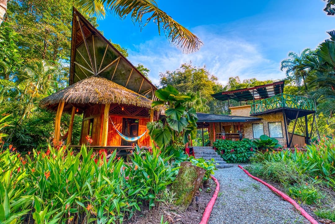 Front-of-house at midday — thatched rancho with hammock strung between hardwood posts, master balcony above